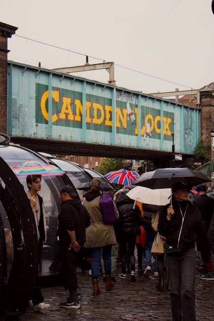 A large, weathered railway bridge with the words 'Camden Lock' painted in bold yellow letters against a blue-green metal surface, spanning over a busy street intersection in Camden Town, London. The b