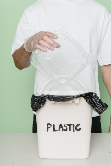 A man with a neutral expression standing behind a table or counter, sorting waste materials into separate recycling bins labeled 'PLASTIC' and 'TRASH,' with a green recycling bin to his left. The man 