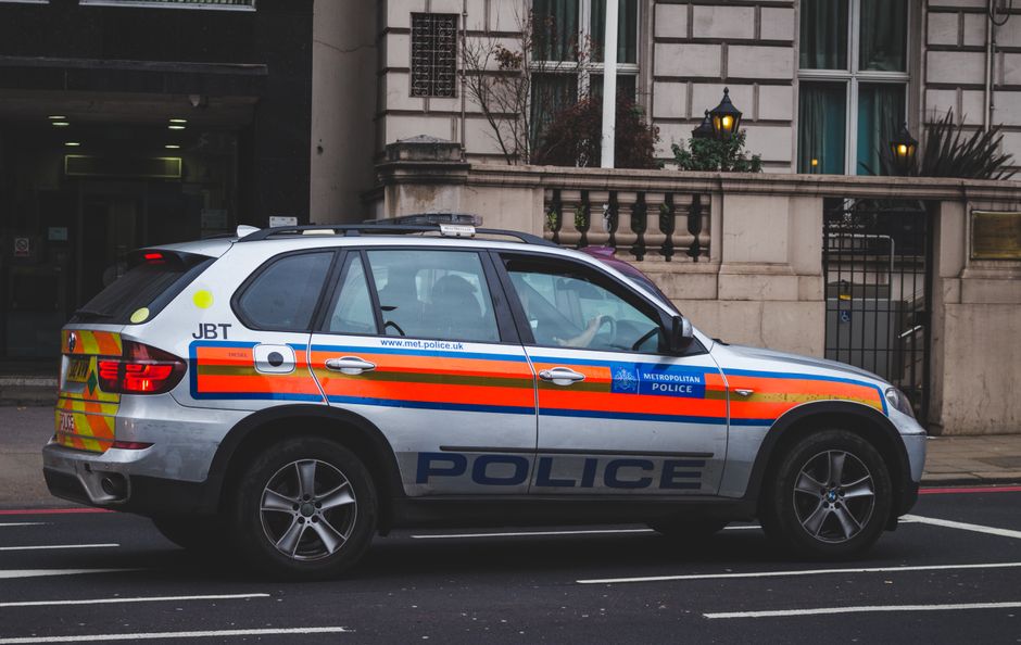 A police vehicle parked on a city street in front of a building with a stone facade, featuring a silver body with orange, blue, and yellow reflective markings, and the word 'POLICE' prominently displa