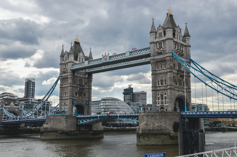 Image showing Tower Bridge over the River Thames in London on a clear day with a bright blue sky and some scattered clouds. In the foreground, there are people sitting and standing along the pavement,