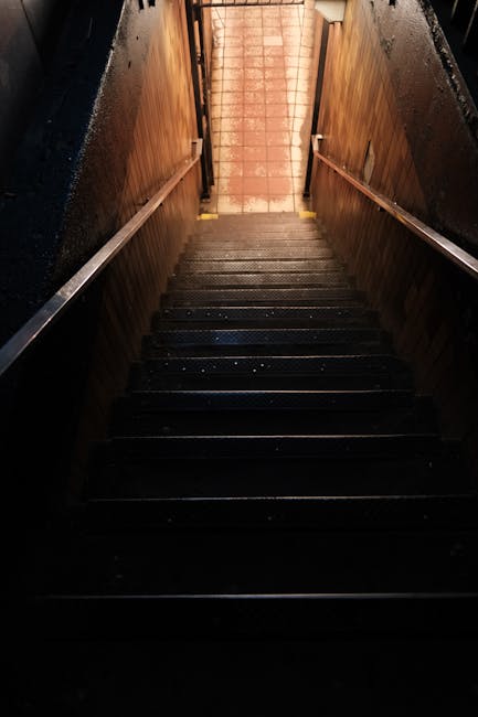 The image shows a narrow alley between two tall buildings in an urban area, with a fire escape staircase attached to the side of the building on the left. The staircase is made of metal with small lan