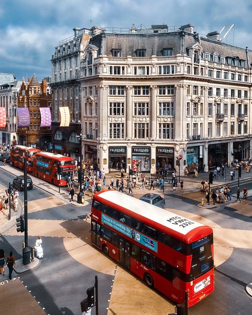 This street scene in London features a red double-decker bus stopping on the road amidst other vehicles, including cars and a bicycle rider. Pedestrians are walking along the pavement adjacent to mult